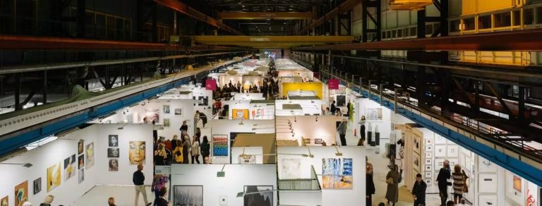 Vista aérea de una nave industrial con stands de una feria de arte y público circulando
