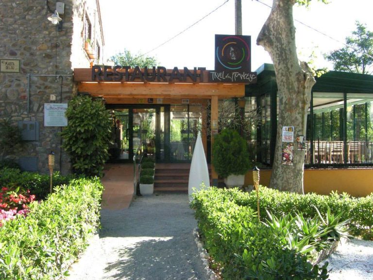 Entrada exterior de un restaurante con pérgola de madera y jardín, sede de la exposición "Art de dos".