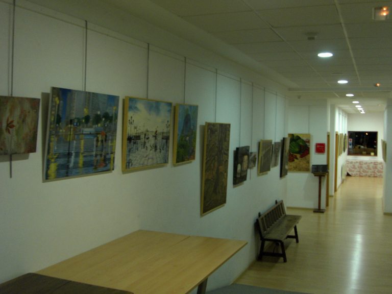 Pasillo de una galería con varios cuadros colgados de hilos desde el techo, una mesa y un banco