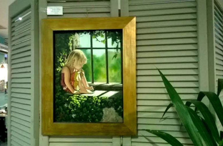 Cuadro de una niña rubia asomada a una ventana entre hojas verdes, colgado sobre pared de listones con una pequeña etiqueta ilegible encima