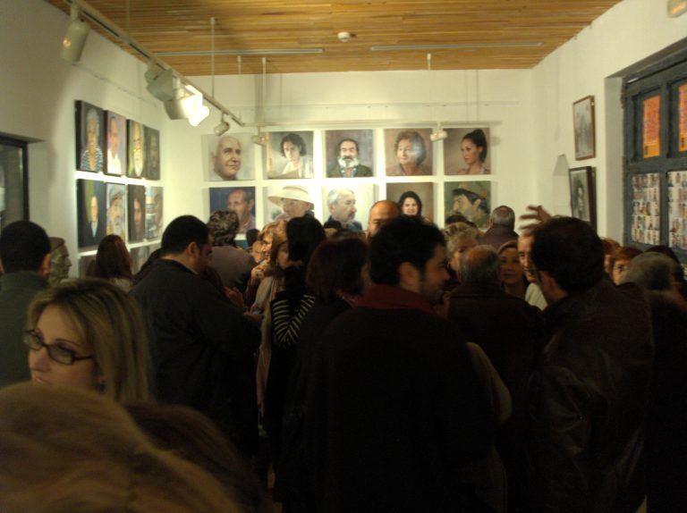 Sala llena de gente con una pared de retratos pintados al fondo.