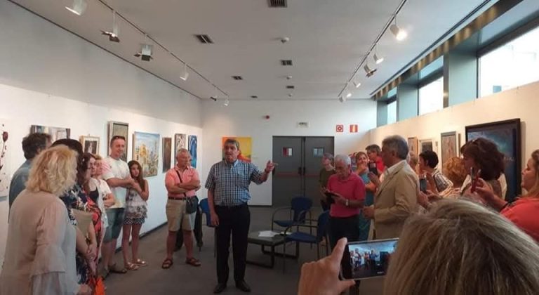 Sala de exposiciones con público escuchando a un hombre hablar, cuadros colgados en las paredes.